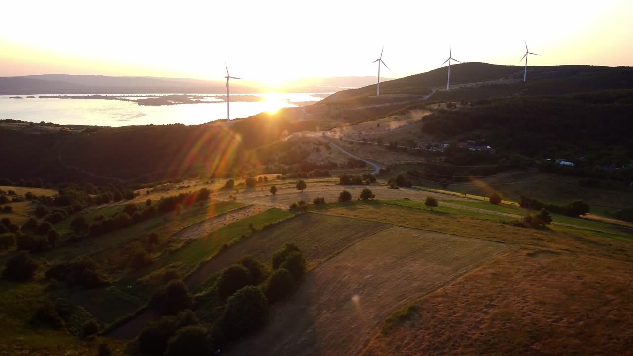 Český Banát-Aerial drone push forward over Romania's rural fields at sunset, wind turbines spin over farmland and hills with lake views.