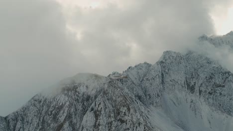 Flying-through-clouds-and-fog-revealing-the-mountain-top-of-the-north-chain-alps-in-austria-and-cabin-covered-surrounded-by-clouds