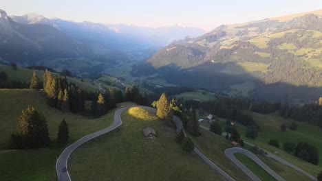 Drone-shot-following-blue-car-driving-on-swiss-mountain-road-in-summer-with-mountains-in-the-background