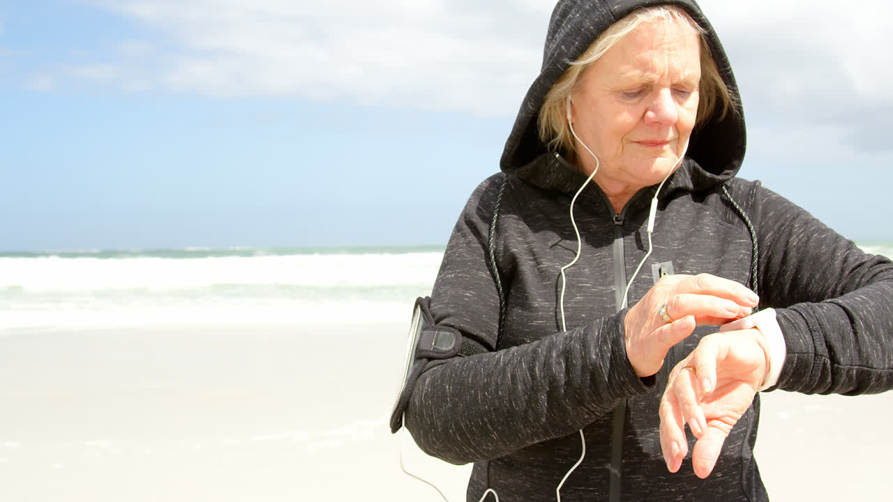 Front view of old caucasian senior woman using smartwatch and listening music on earphones at beach