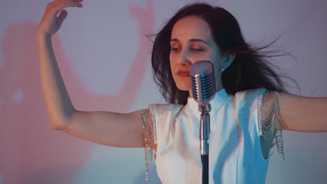 singer in a white gown gracefully moving her hands in the air while performing into a vintage microphone, with hair flicking against a white background accented by soft pink and blue lighting