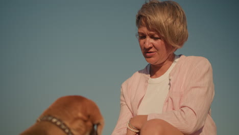 woman in pink shirt standing outdoors with blue sky background, interacting with her brown dog in a calm and caring manner, preparing to feed her pet