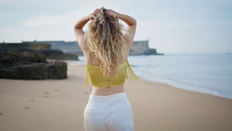 serene woman strolling beach touching curly hair back view. girl walking sand