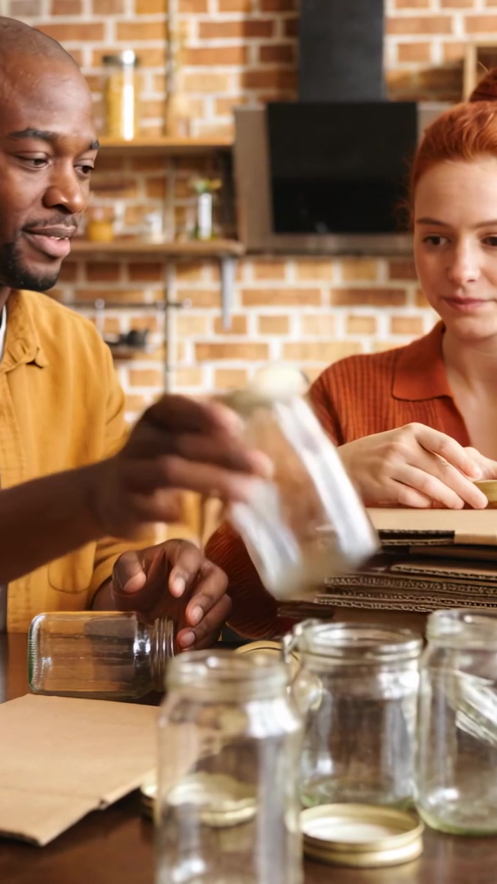 Video verticale: Selezione di barattoli, uomo in giallo, donna in arancione che smistano coperchi e fogli in cucina per la conservazione.