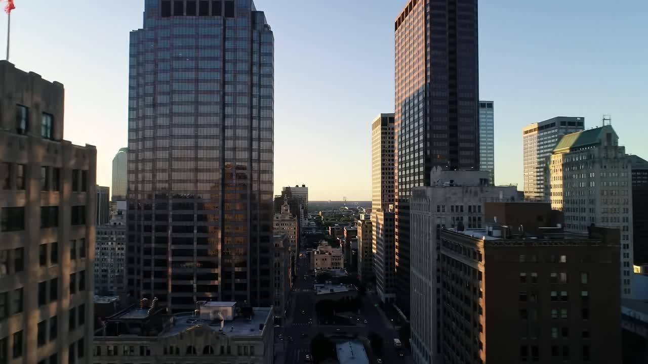 The skyline showcases tall buildings reflecting the warm colors of sunset. This urban setting highlights the blend of modern architecture and a vibrant city atmosphere during evening hours.