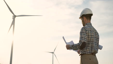 Rückansicht-Des-Kaukasischen-Ingenieurs,-Der-Einen-Helm-Trägt-Und-Einige-Blaupausen-An-Der-Windstation-Für-Erneuerbare-Energien-Anschaut