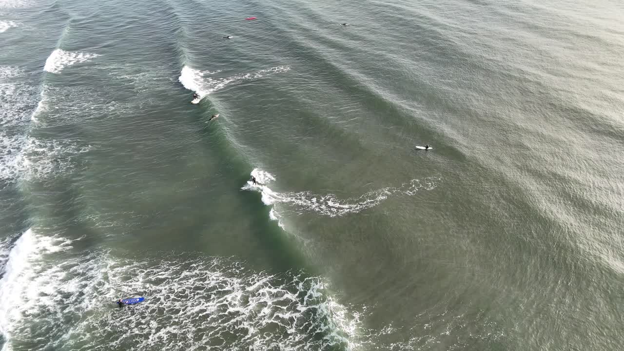 Aerial footage captures multiple surfers paddling and riding waves in the gentle morning light at West Sands Beach, St Andrews, Scotland. Smooth camera movement follows the action