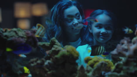 mother-with-daughter-looking-at-tropical-fish-in-aquarium-tank-curious-little-girl-watching-marine-animals-swimming-in-corel-reef-habitat-child-learning-about-sea-life-with-mom