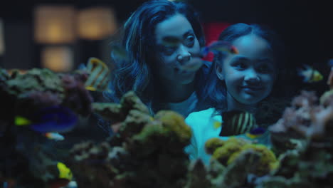 mother with daughter looking at tropical fish in aquarium tank curious little girl watching marine animals swimming in corel reef habitat child learning about sea life with mom