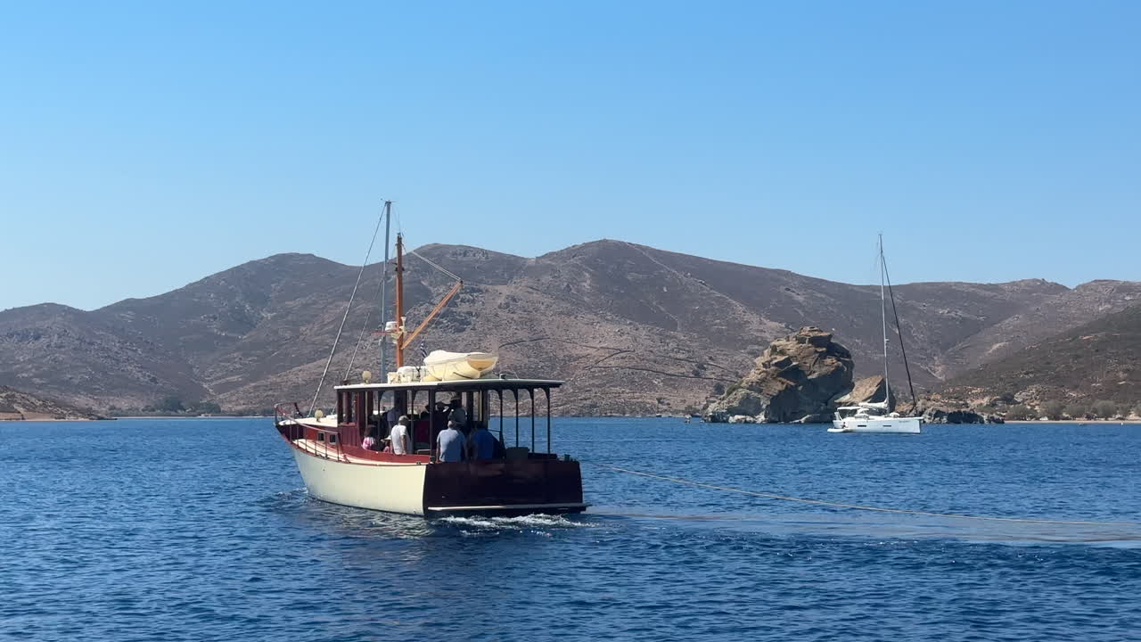 Greece, Patmos Island, Grikos Bay, a beautiful white classical wooden trawler is in motion and leaving the bay. Trawler and the sailboat move together towards the same direction.