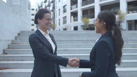 side view of happy female business partners meeting outside, shaking hands, smiling and talking together