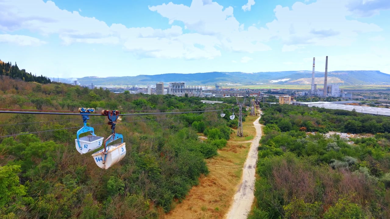 Passeio de teleférico sobre um vale verde. Uma pessoa viaja em um teleférico sobre árvores e caminhos em um vale com fábricas ao longe em um dia claro.