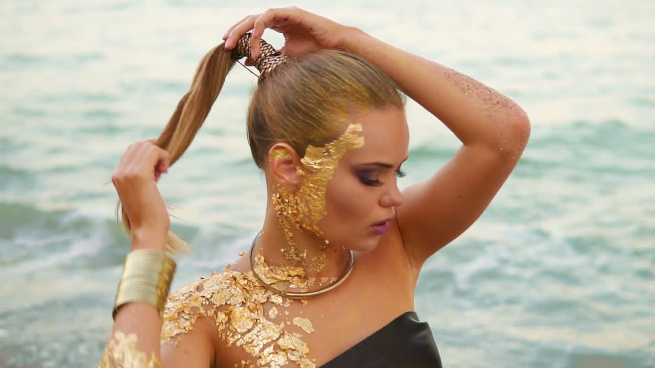 красивая женщина с профессиональным золотым макияжем смотрит в камеру и флиртует с зрителем, касаясь ее волос. сексуально