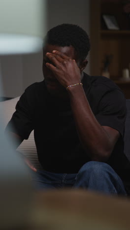 vertical video of unhappy and depressed young man sitting on sofa at home looking anxious and worried resting head on hand 10