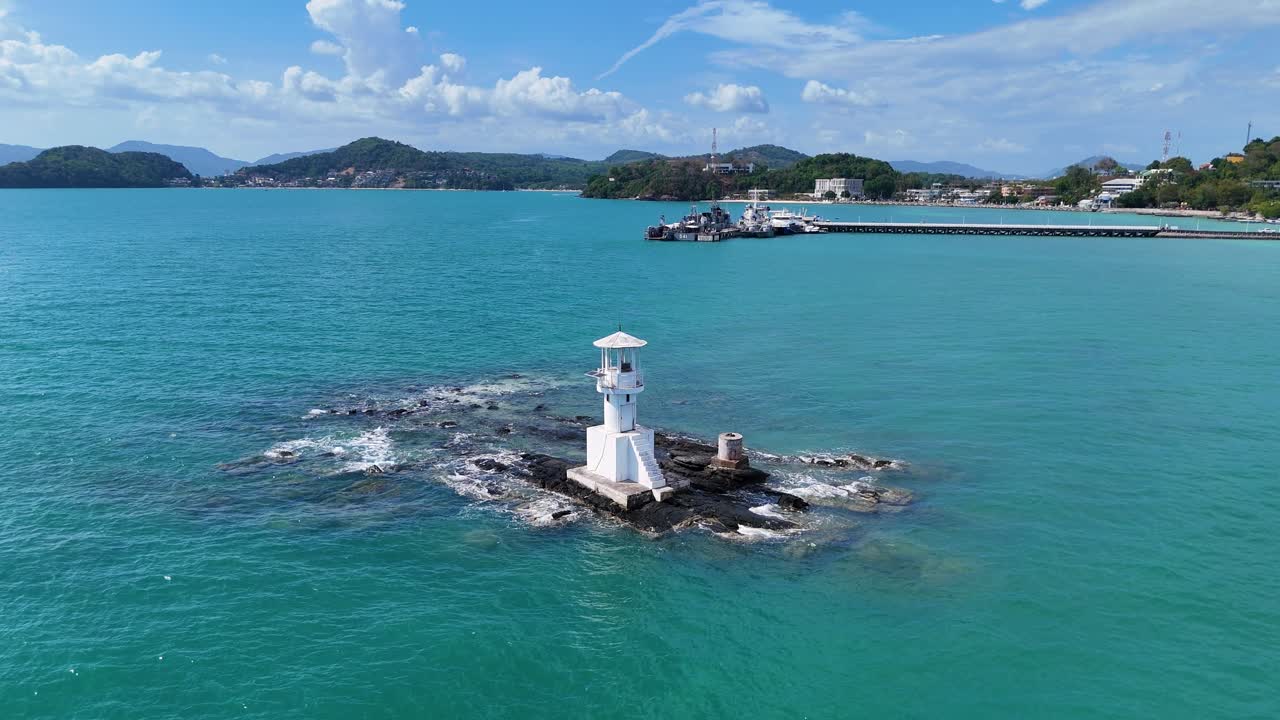 Drone footage captures a serene lighthouse on rocky outcrop in Phuket, Thailand, surrounded by turquoise waters under clear skies