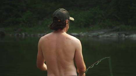 Parte-Trasera-De-Un-Joven-Adulto-En-Topless-Pescando-Con-Mosca,-Lanzando-Hilo-De-Pescar-Al-Lago