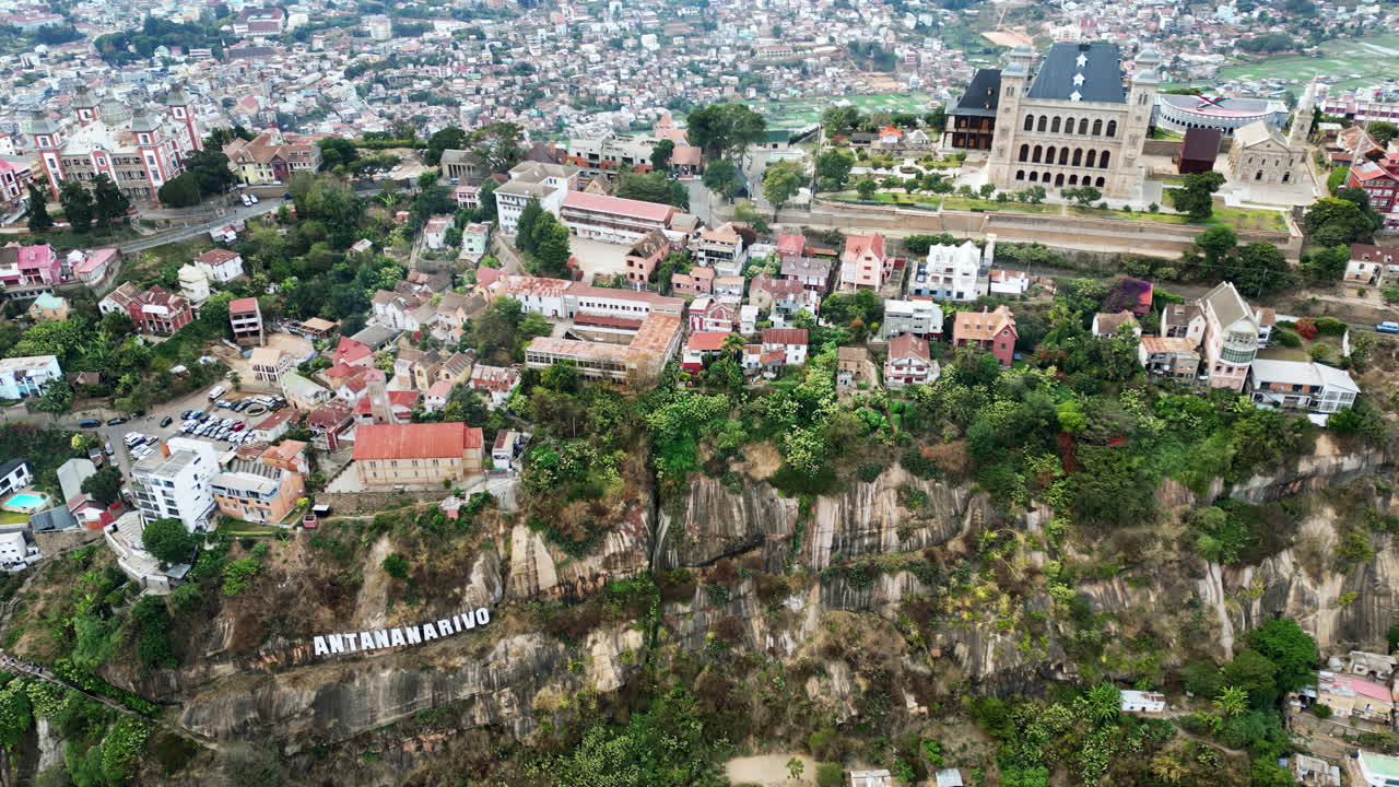 Aerial drone view above Antananarivo with the city skyline spread across surrounding hills, showing dense urban neighborhoods and the large historic palace complex overlooking Madagascar's capital