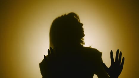 silhouette of woman emerges from smoke and touches glass with her palms - silhouette of woman emerges from smoke and touches glass with her palms. woman examines glass and slides her hands over it with interest. model poses in dark studio with yellow soft lights in background. close up