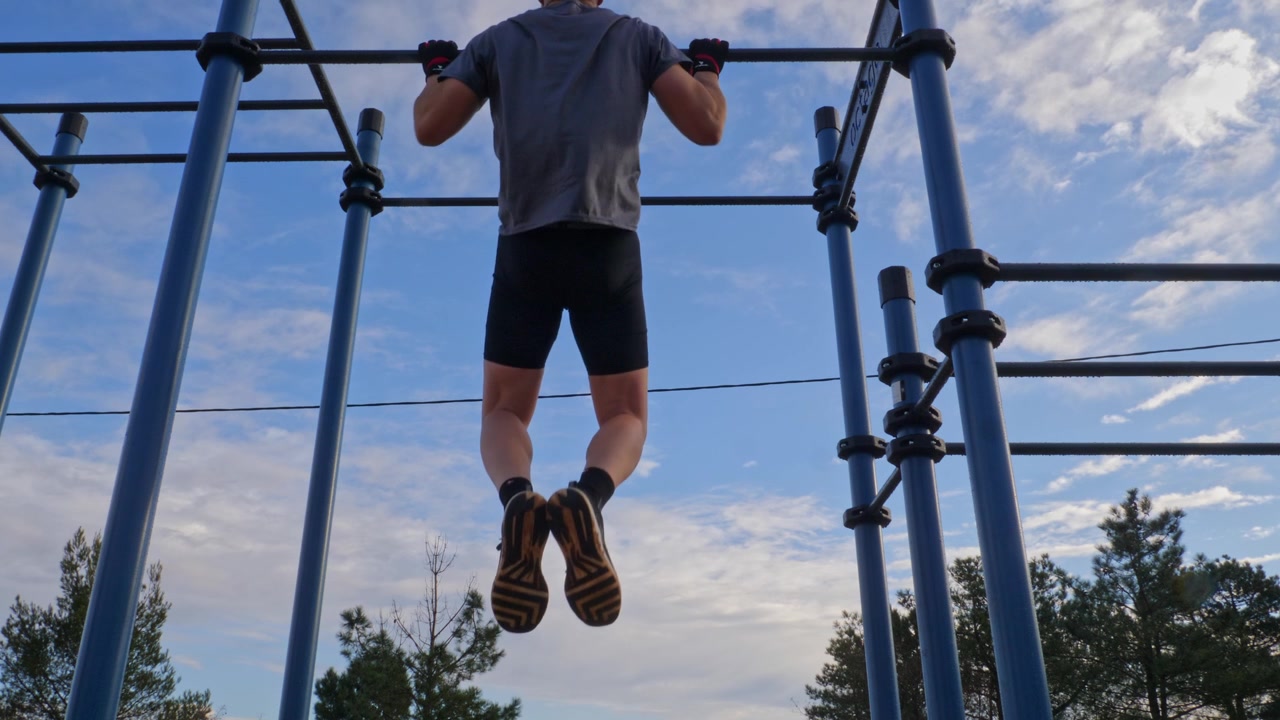 Cena Estática de um Homem em Forma Fazendo Barras em um Parque Público. O Atleta Veste Roupa Esportiva Preta e Cinza com Luvas Sem Dedos. Conceito de Força e Fitness ao Ar Livre