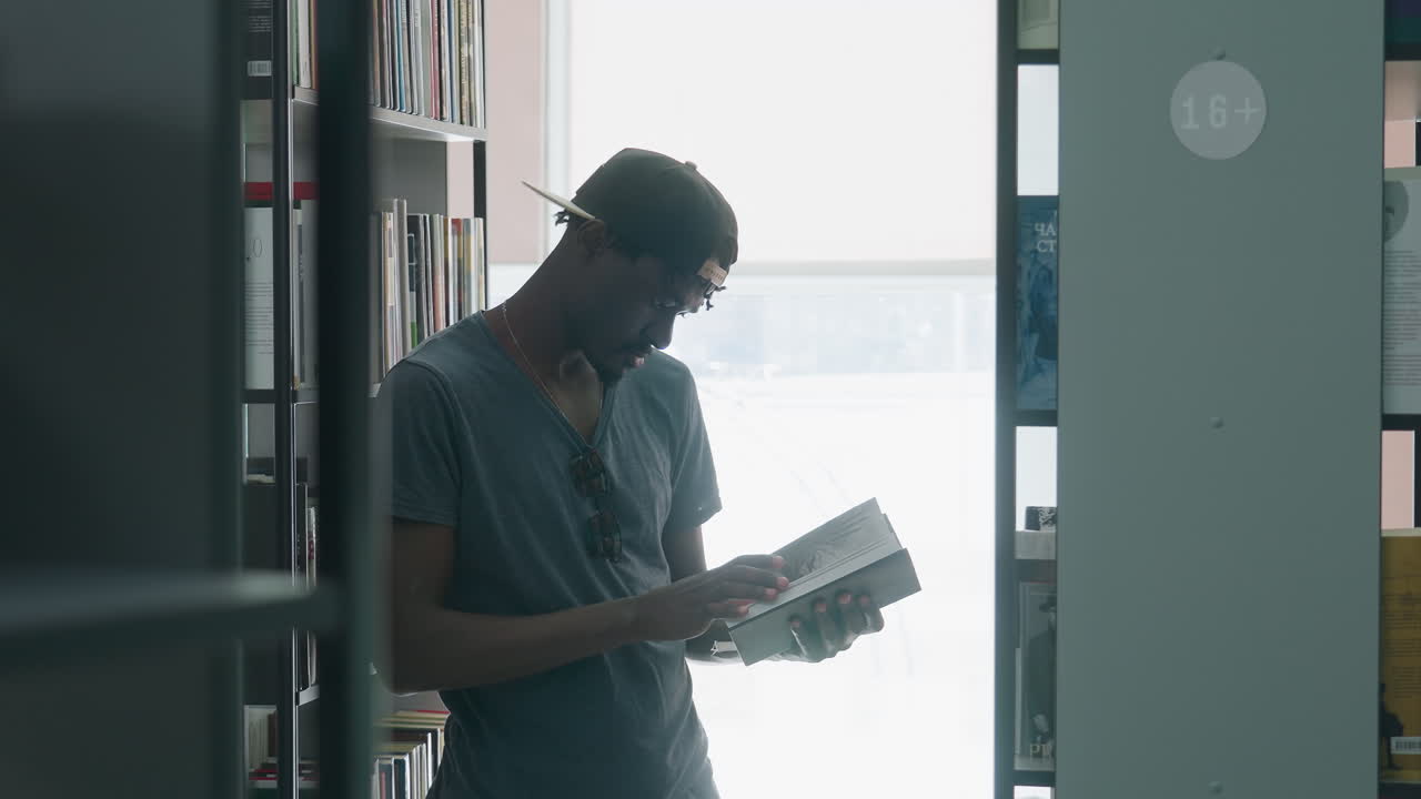 Side view of young man in cap and t-shirt standing between bookshelves in bright library, holding open book with one hand while turning pages with other