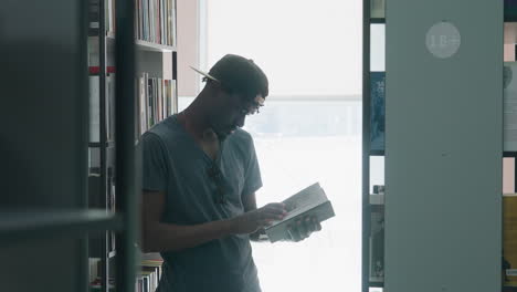 side view of young man in cap and t-shirt standing between bookshelves in bright library, holding open book with one hand while turning pages with other