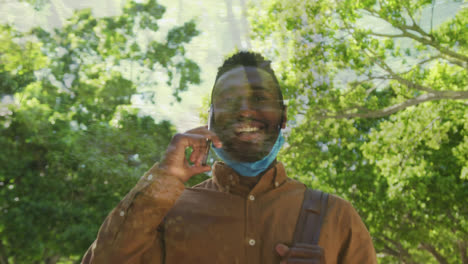 Forest-path-against-with-african-american-man-lowered-face-mask-talking-on-smartphone-while-walking