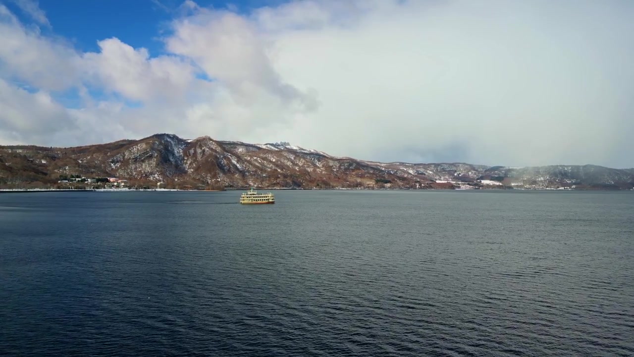 Vakkert dronebilde av en fjern turistbåt som seiler over en bred blå innsjø med snødekte vulkanske fjell under en skyet himmel.