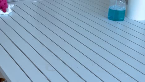 a person wearing pink gloves cleans a white table using a cloth and cleaning solution in a bright setting
