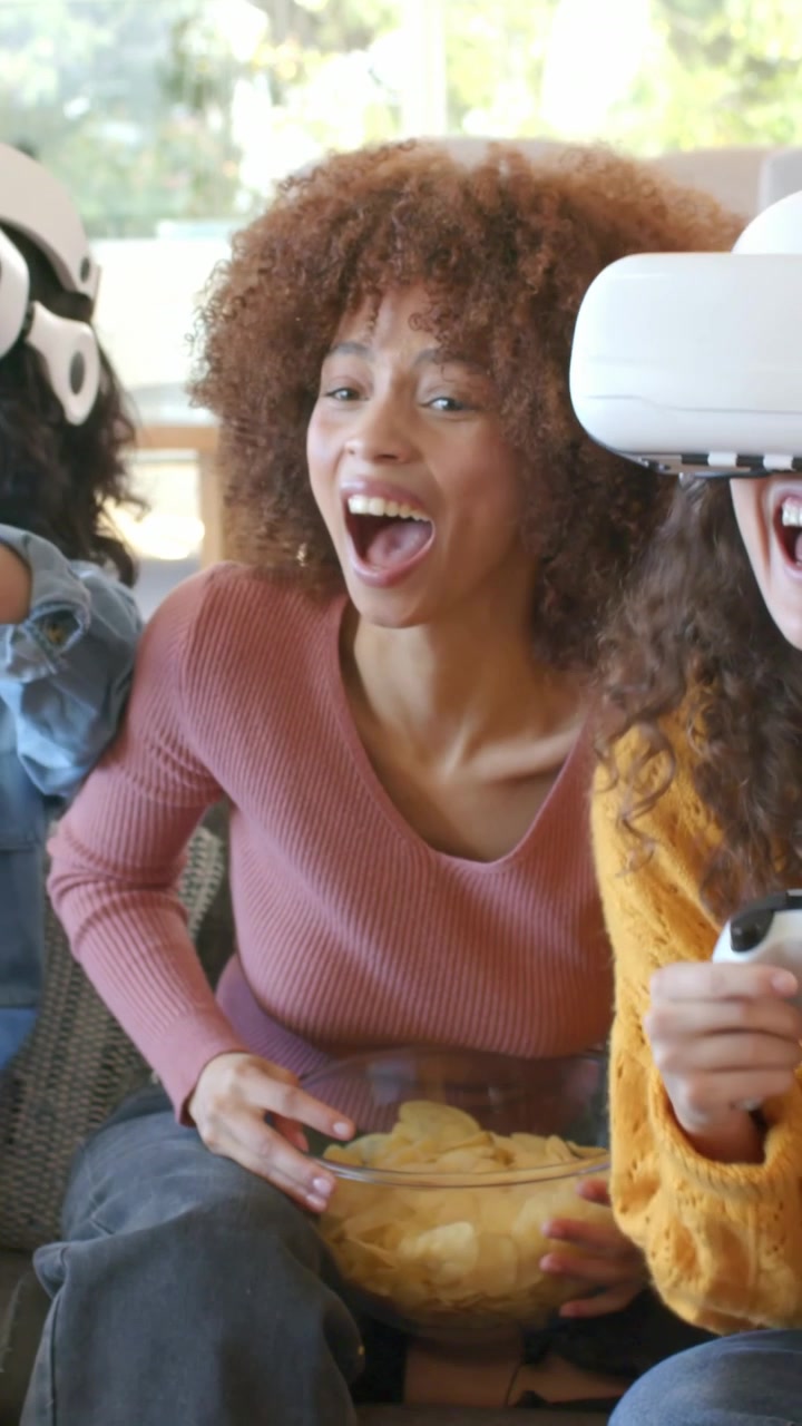 Video vertical: Mujer sonriente con top rosa reaccionando a amiga poniéndose equipo de realidad virtual en el sofá, sosteniendo patatas fritas