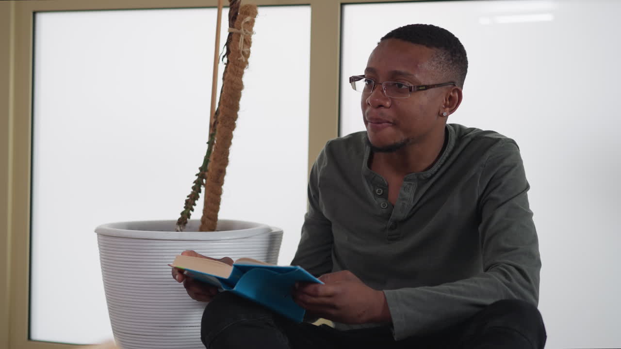 hombre con libro se sienta en el suelo contra la ventana. joven lector afroamericano comparte pensamientos sobre la literatura en la librería. estudiante con libro de texto en la universidad