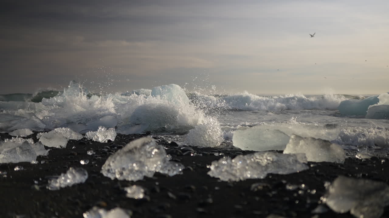Waves crash on glacial ice on black sand beach under moody sky at Diamond Beach, Iceland