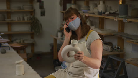 female caucasian potter wearing face mask and apron holding pot and talking on smartphone at pottery