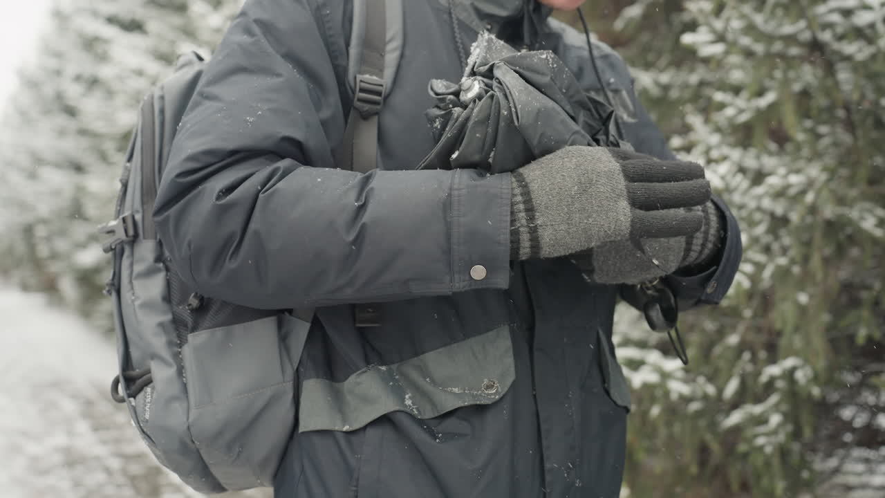 Mann mit Rucksack entfaltet Regenschirm neben schneebedeckten Kiefern, graue Strickhandschuhe bearbeiten Speichen und Schirmdach, Wassertropfen und leichte Schneeflocken, Fokus auf Hände und Ausrüstung, während Wanderer sich auf raueres Wetter vorbereitet.