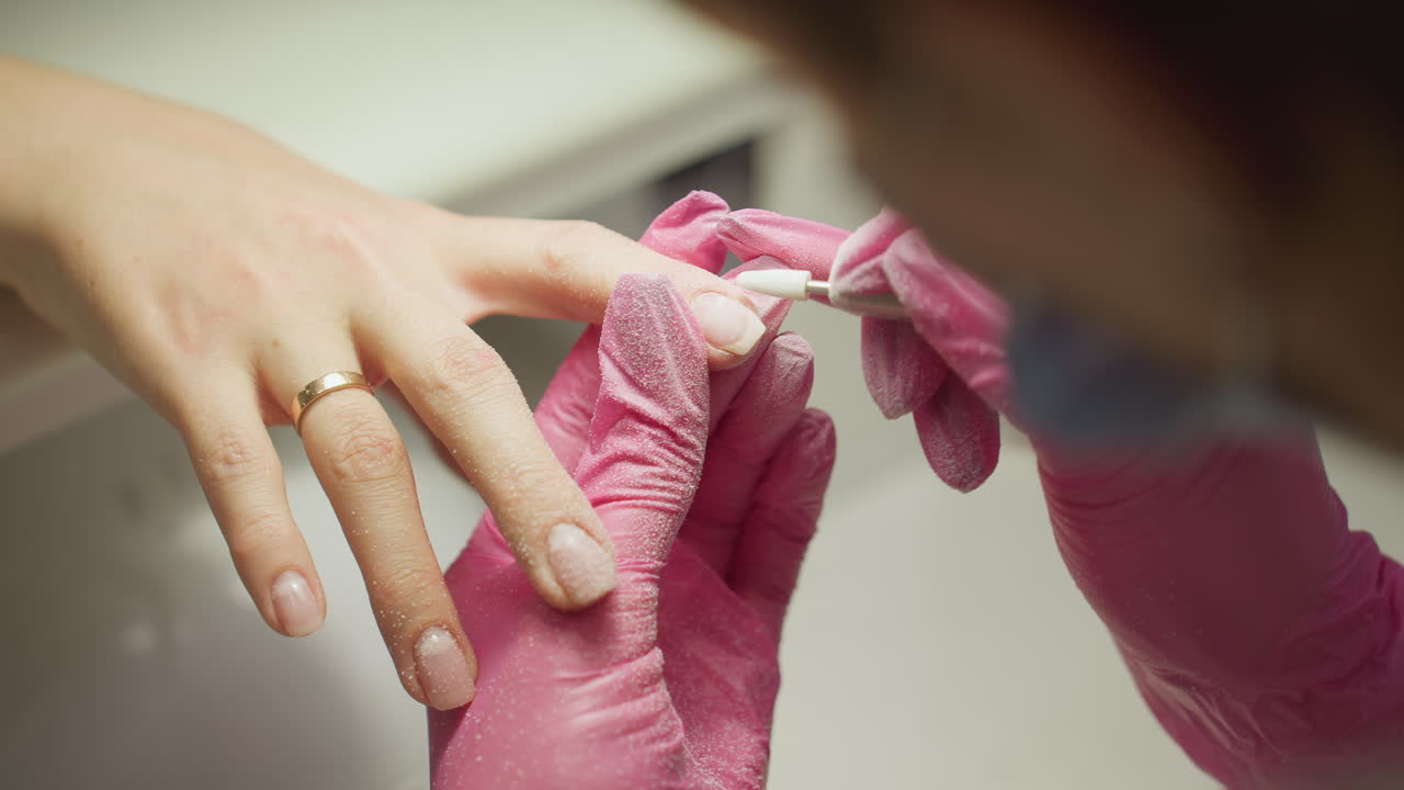 Close-up of nail technician in pink gloves using electric file on client nail during manicure, with dust particles covering gloves as nail is shaped and carefully handles client hand for precise work