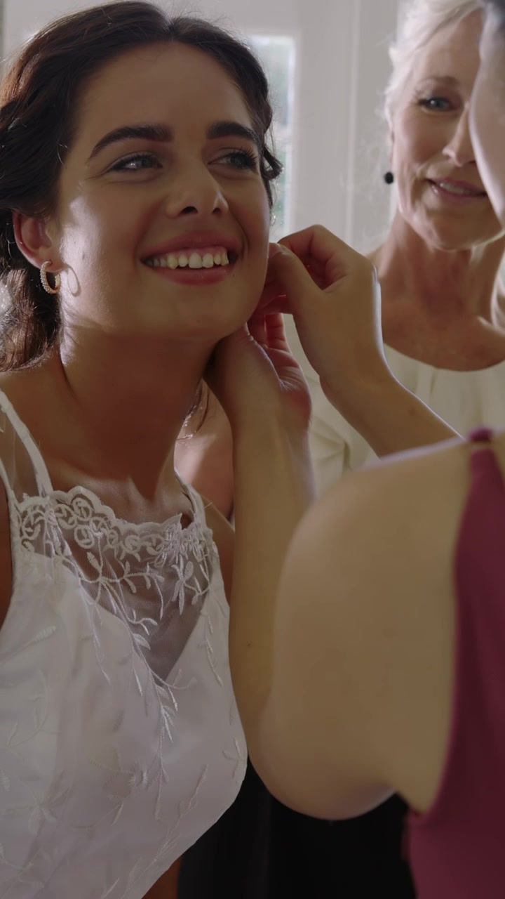 Vertikal video: Brud, eldre mor og asiatisk venninne fikser øredobber mens spray gnistrer ved vinduet til bryllup