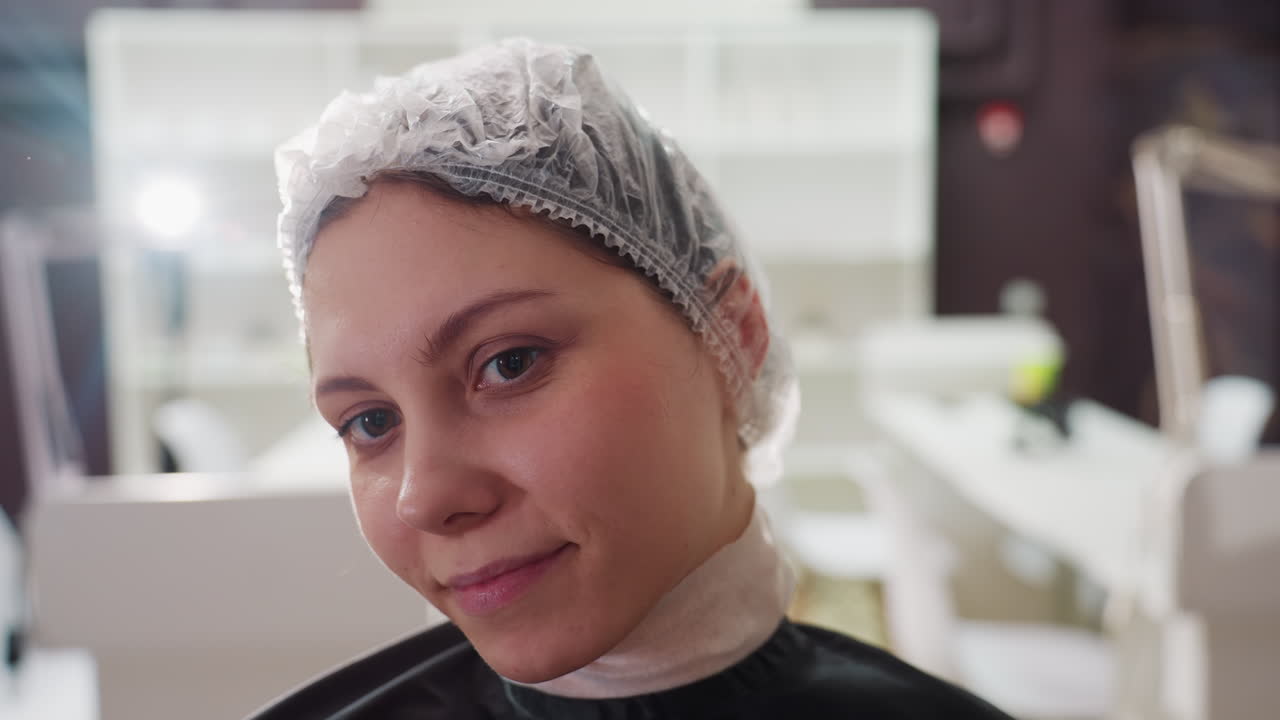 Primer plano de una mujer caucásica en un salón de belleza con gorro mientras le preparan un tratamiento capilar químico; esteticista preparando el cuero cabelludo, gorro y capa desechables, expresión tranquila y confiada, iluminación clínica suave, salón.