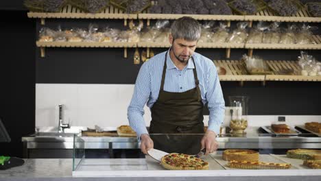 Young-handsome-man-vendor-in-the-apron-bringing-fresh-pies-to-the-counter-in-the-bakery-shop.-Indoors