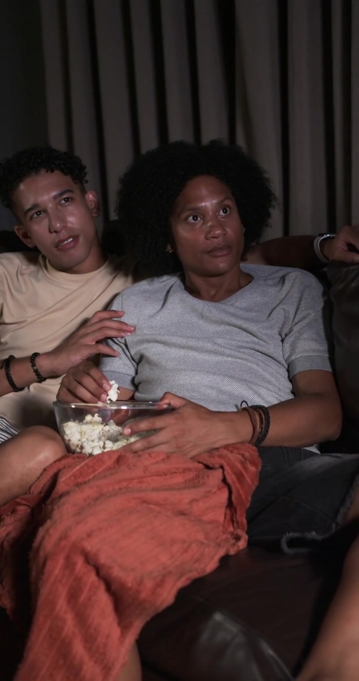 Video verticale: Coppia illuminata dalla luce della TV che guarda a casa, condividendo una ciotola di popcorn sotto una coperta arancione.