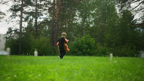 Young boy dressed in black outfit walks across green grassy field carrying soft teddy bear, surrounded by tall trees with distant buildings and fence partially visible in background on a cloudy day