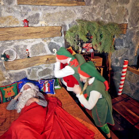 santa claus is sleeping in specially decorated room in his bed. hand under cheek. elves are ringing bell and waving hands near his face. christmas spirit concept close up video. video from above.