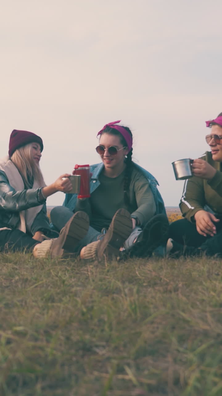 happy young women tourists drink alcohol from cups sitting on grass by tent against clear evening sky low angle shot