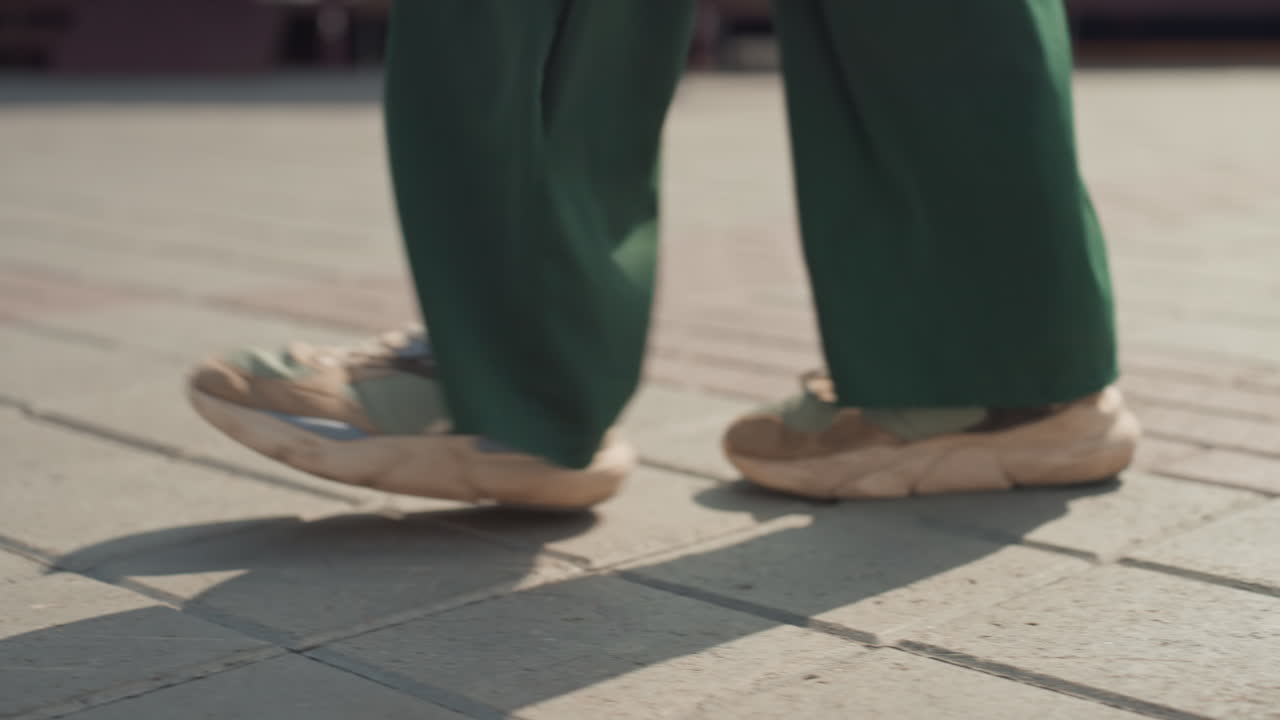 Kaldırımda yürüyen bacakların yakın çekimi, yeşil pantolon ve bej spor ayakkabılarla hafif adımlar, şehir meydanı, yumuşak güneş ışığı, uzun gölgeler, dokulu kaldırım taşları, yavaş çekimde yakalanan gündelik işe gidip gelme hareketi