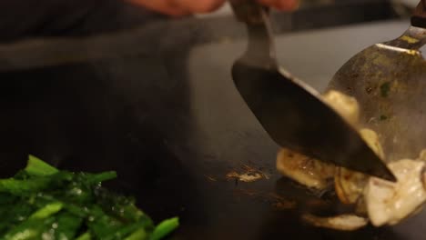 close-up of oysters and greens being stir-fried on a hot griddle with a metal spatula.