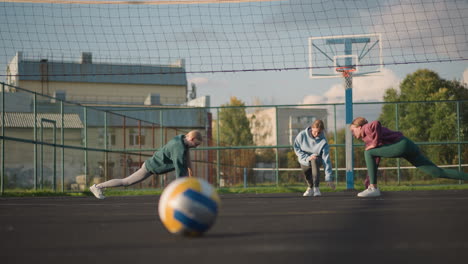 close-up of volleyball on ground with blurred background of athletes stretching and exercising outdoors, volleyball net and sports court visible with distant buildings under clear sky