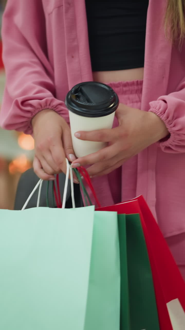 dama con una camiseta negra bajo un vestido rosa recoge su taza de café de la mesa, sosteniendo varias bolsas de compras, preparándose para irse, con iluminación suave de color y fondo borroso