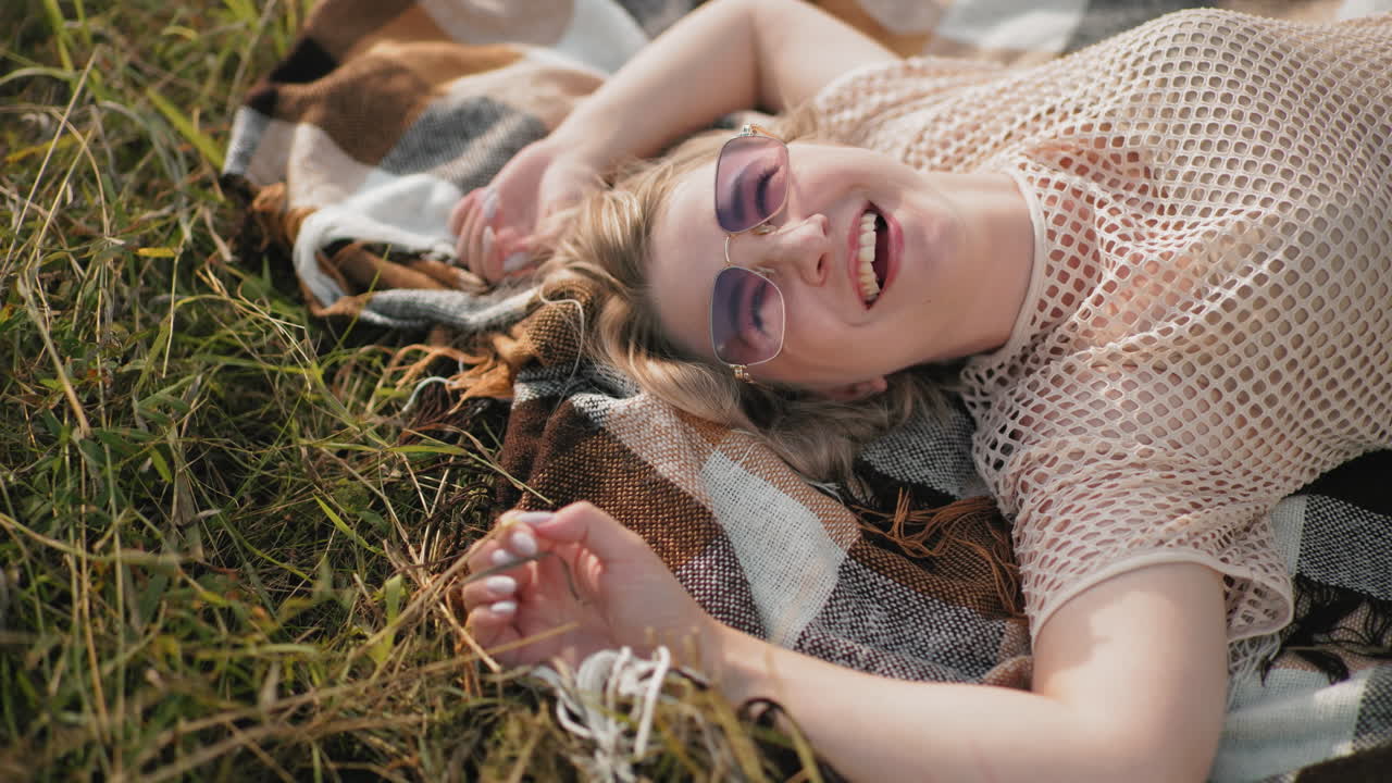 mujer feliz con camiseta y gafas de sol acostada en la sábana al aire libre, sonriendo y tocando la hierba, momento de relajación en la naturaleza con cálida luz solar y atmósfera pacífica