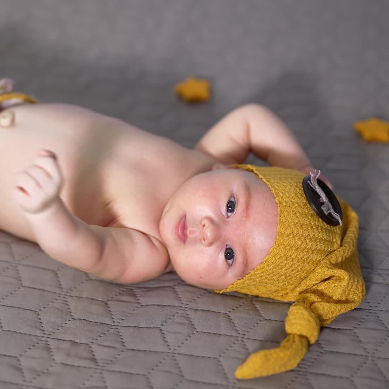 Toddler in a yellow cap with button on the bed. Baby in a funny costume tossing arms and feet. Nice kid on a grey background
