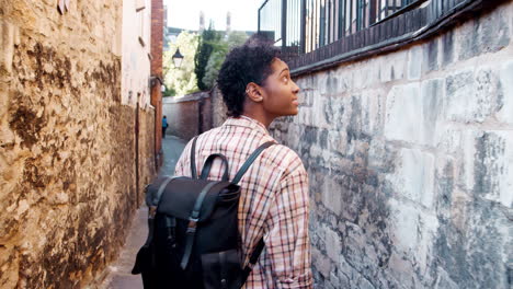 young black woman with a backpack walking in a narrow alleyway between stone walls, back view, follow shot