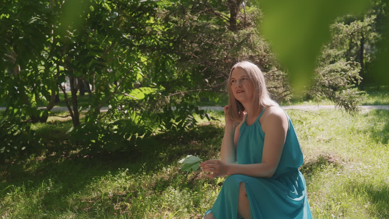 Mujer descansando al aire libre. Mujer relajándose en el césped bajo árboles soleados. Mujer sentada en un parque en un día soleado de verano. Dama vestida de turquesa descansando tranquilamente entre la hierba sombreada y la luz del sol.