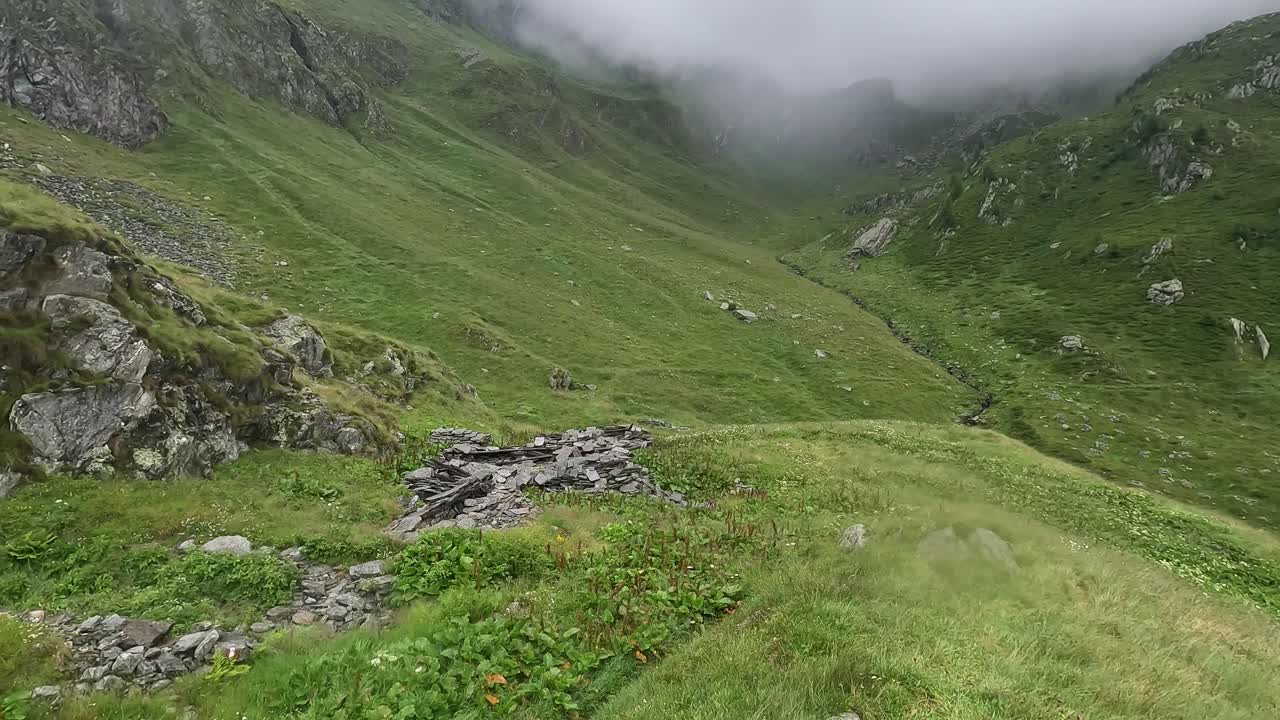 Fog drifts over a lush alpine valley with green slopes and rocky terrain in Val Cairasca, Alpe Veglia